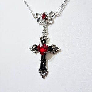 Puede incluir: Collar plateado con un colgante de cruz g&oacute;tica con una gema roja en forma de coraz&oacute;n en el centro. La cruz es negra con detalles ornamentados. Una gema roja m&aacute;s peque&ntilde;a est&aacute; colocada encima. La cadena es delicada. El colgante mide unos 5 cm de largo.