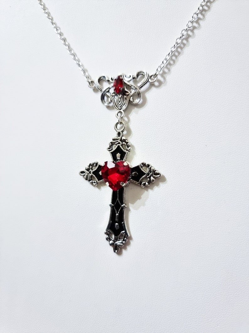 Puede incluir: Collar plateado con un colgante de cruz g&oacute;tica con una gema roja en forma de coraz&oacute;n en el centro. La cruz es negra con detalles ornamentados. Una gema roja m&aacute;s peque&ntilde;a est&aacute; colocada encima. La cadena es delicada. El colgante mide unos 5 cm de largo.