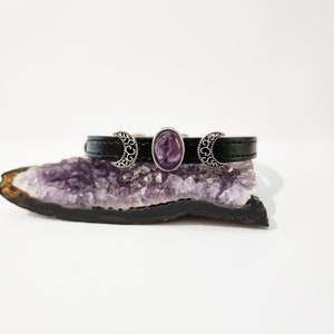 May include: A black leather bracelet with three silver crescent moon charms and a purple amethyst stone in the center.