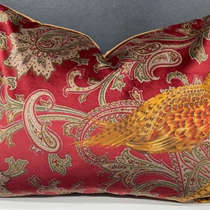 May include: A decorative throw pillow with a red paisley pattern and a detailed pheasant illustration in shades of orange, yellow, and brown. The pillow has a gold trim and is designed for home decor.