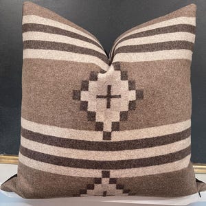 May include: A square throw pillow featuring a geometric pattern in shades of brown, tan, and cream. The design includes horizontal stripes and a central diamond-shaped motif with a cross symbol, all rendered in a textured, woven fabric finish.