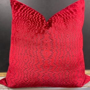 May include: A plush, square decorative pillow in a vibrant red hue. The pillow features a detailed, textured pattern across its surface. The fabric appears to be a soft velvet or similar material, adding to its luxurious appearance. The pillow is designed for home decor.