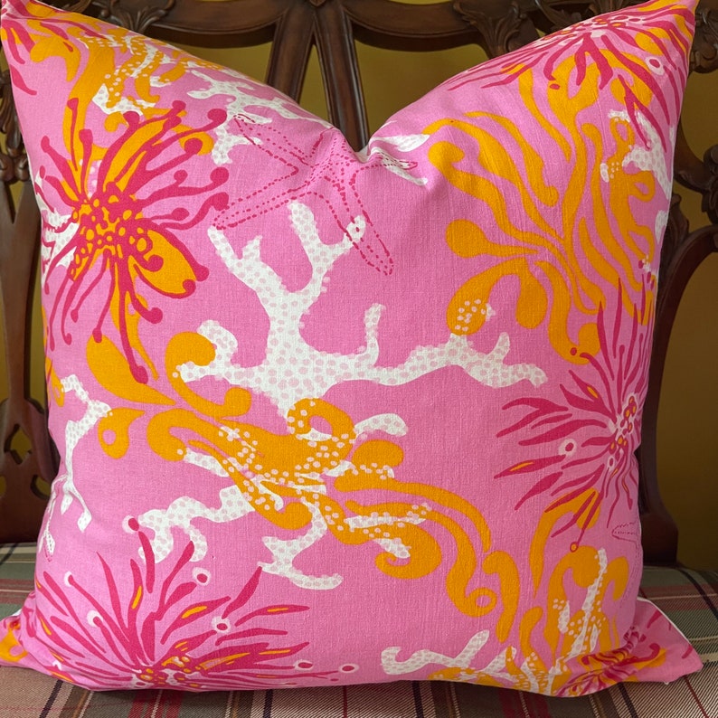 May include: Pink pillow with a white and yellow coral reef design. The pillow features a repeating pattern of coral, starfish, and sea anemones.