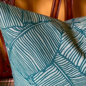 May include: A teal blue throw pillow with a white leaf pattern. The pillow is made of a textured fabric and has a subtle sheen.
