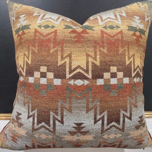 May include: A square throw pillow with a geometric pattern in shades of brown, orange, and gray. The pillow has a woven texture and a fringe border.