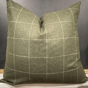 May include: A square, olive green throw pillow with a white grid pattern. The pillow has a soft, textured appearance and is likely made of a woven fabric. The pillow is a decorative item for a bed or sofa.
