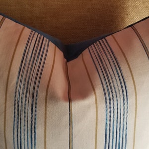 May include: A striped pillow with blue and gold stripes on a white background. The pillow has a blue border and is sitting on a chair.