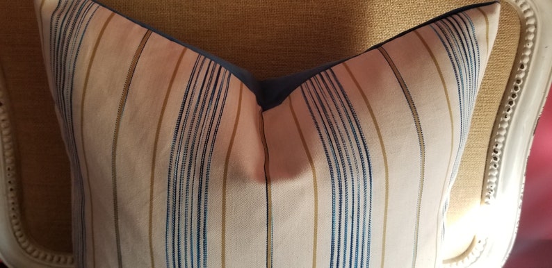 May include: A striped pillow with blue and gold stripes on a white background. The pillow has a blue border and is sitting on a chair.