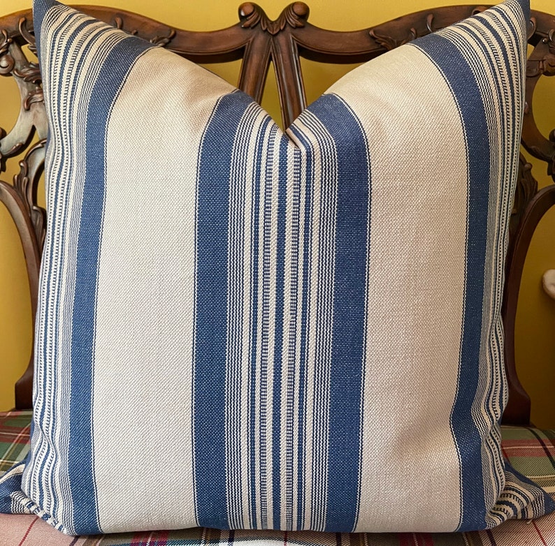 Puede incluir: Una almohada de tiro a rayas azules y blancas con una textura tejida sutil. La almohada se muestra en una silla de madera con una tela a cuadros.
