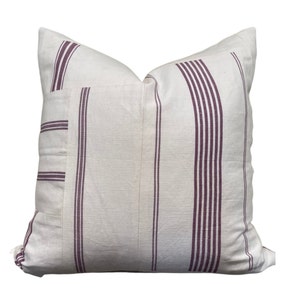 May include: A square pillow with a cream-colored background and vertical burgundy stripes. The pillow has a sewn-on patch with matching stripes. The pillow is made of a textured fabric.