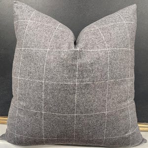 May include: A square gray decorative pillow with a white grid pattern. The pillow has a soft, textured appearance and is likely made of a woven fabric. The pillow is designed for home decor.