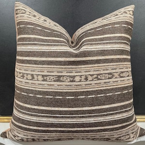 May include: A square throw pillow featuring a woven pattern of horizontal stripes in shades of dark brown, tan, and cream. The design includes alternating bands of solid lines and intricate, repeating geometric motifs across the entire surface.