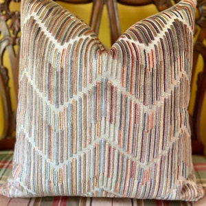 May include: A decorative throw pillow with a geometric pattern in shades of beige, brown, orange, and red. The pillow has a textured, woven fabric with a subtle sheen.