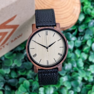 Pu&ograve; includere: Un orologio da polso in legno con un cinturino in pelle nera. Il quadrante dell'orologio &egrave; bianco con indici orari in numeri romani neri e lancette nere. L'orologio &egrave; su uno sfondo di foglie verdi.