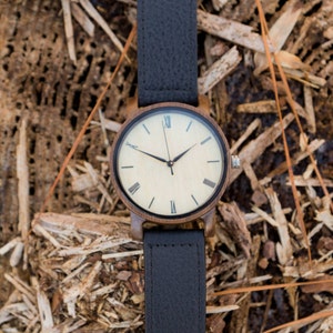 Pu&ograve; includere: Un orologio da polso in legno con un cinturino in pelle nera. Il quadrante dell'orologio &egrave; bianco con indici orari in numeri romani neri e lancette nere.