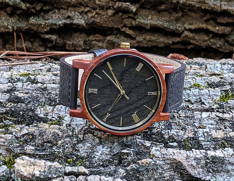 May include: A wooden wristwatch with a black face and gold Roman numeral hour markers. The watch has a brown leather strap.