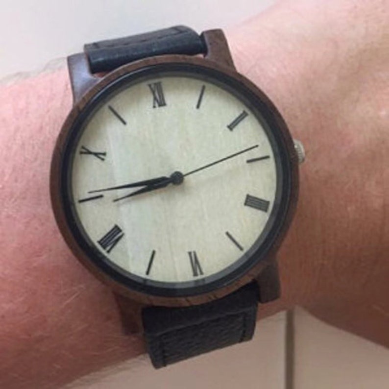 Pu&ograve; includere: Un orologio da polso in legno marrone con cinturino in pelle nera e quadrante bianco con indici orari in numeri romani.