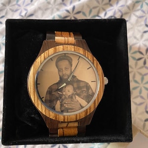 May include: Wooden wristwatch with a light brown and dark brown striped design. The watch face features a black and white photograph of a person holding a baby. The watch is presented in a black box.