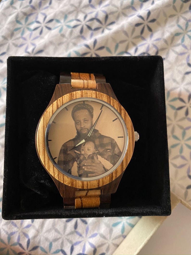 May include: Wooden wristwatch with a light brown and dark brown striped design. The watch face features a black and white photograph of a person holding a baby. The watch is presented in a black box.