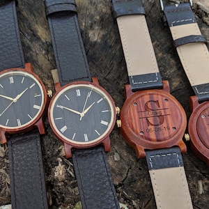 May include: Four wooden wristwatches with black leather straps. Two watches have black faces with gold Roman numeral markings. Two watches have engraved wooden backs with names and dates.