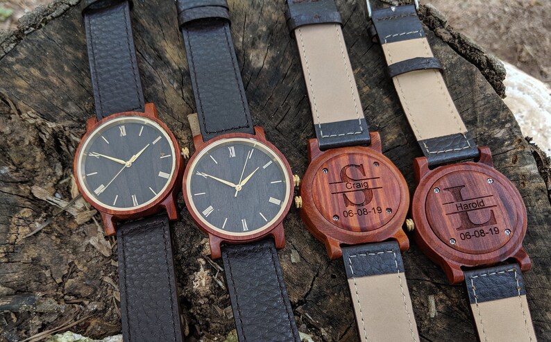 May include: Four wooden wristwatches with black leather straps. Two watches have black faces with gold Roman numeral markings. Two watches have engraved wooden backs with names and dates.