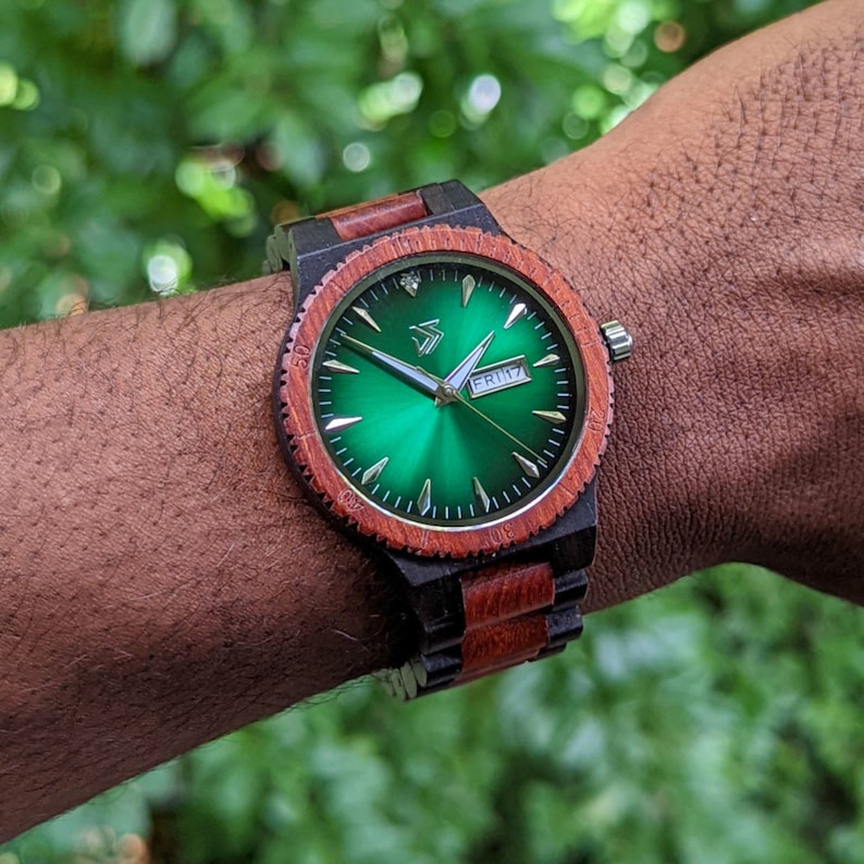 Peut inclure: Une montre-bracelet en bois avec un bracelet noir et rouge. Le cadran de la montre est vert avec des aiguilles et des marqueurs argent&eacute;s. La fen&ecirc;tre de la date affiche "FRI 17".