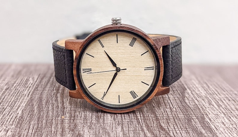 Pu&ograve; includere: Un orologio da polso in legno con cinturino in pelle nera e quadrante bianco con indici orari in numeri romani.