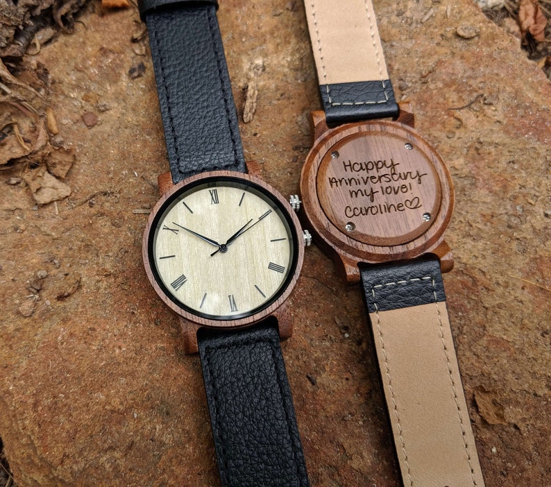 Pu&ograve; includere: Due orologi da polso in legno con cinturini in pelle nera. Un quadrante dell'orologio ha i numeri romani e l'altro ha un messaggio inciso che dice "Happy Anniversary my love! Caroline".