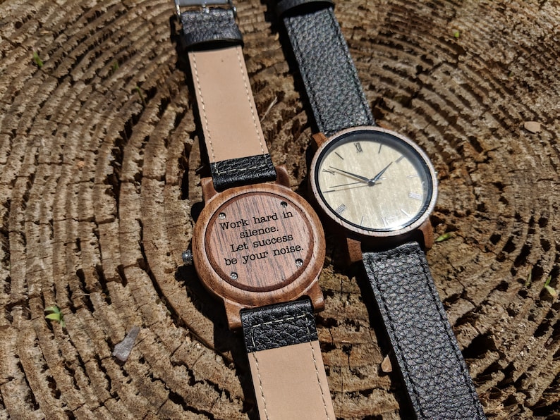 Pu&ograve; includere: Due orologi da polso in legno con cinturini in pelle nera. Un quadrante dell'orologio ha uno sfondo bianco con indici orari romani neri e lancette nere. L'altro quadrante dell'orologio ha uno sfondo in legno con il testo inciso "Lavorare sodo nel silenzio. Lascia che il successo sia il tuo rumore."
