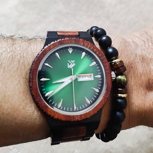 Peut inclure: Une montre-bracelet en bois avec un cadran vert et des aiguilles noires. La montre a un bracelet en bois brun et noir. La montre est port&eacute;e au poignet d'une personne avec un bracelet en perles noires.