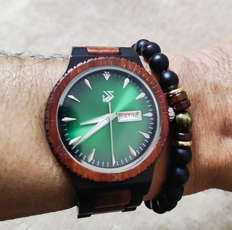 Peut inclure: Une montre-bracelet en bois avec un cadran vert et des aiguilles noires. La montre a un bracelet en bois brun et noir. La montre est port&eacute;e au poignet d'une personne avec un bracelet en perles noires.