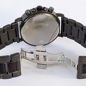 May include: A dark gray chronograph watch with a matching metal band. The watch features a round face with multiple dials and buttons. The band is open, revealing the clasp. The watch is a stylish accessory.