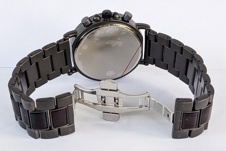May include: A dark gray chronograph watch with a matching metal band. The watch features a round face with multiple dials and buttons. The band is open, revealing the clasp. The watch is a stylish accessory.