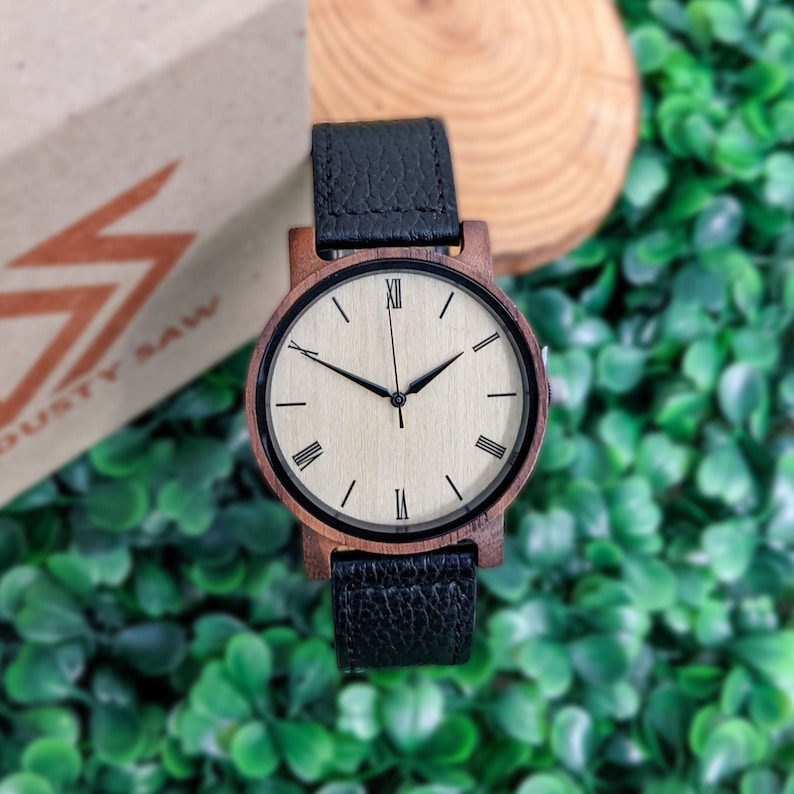 Pu&ograve; includere: Un orologio da polso in legno con un cinturino in pelle nera. Il quadrante dell'orologio &egrave; bianco con indici orari in numeri romani neri e lancette nere. L'orologio &egrave; su uno sfondo di foglie verdi.