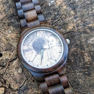 May include: A wooden wristwatch with a dark brown finish. The watch face displays a black and white photograph of two people. The watch band is made of matching wood links. The watch is resting on a weathered wooden surface.