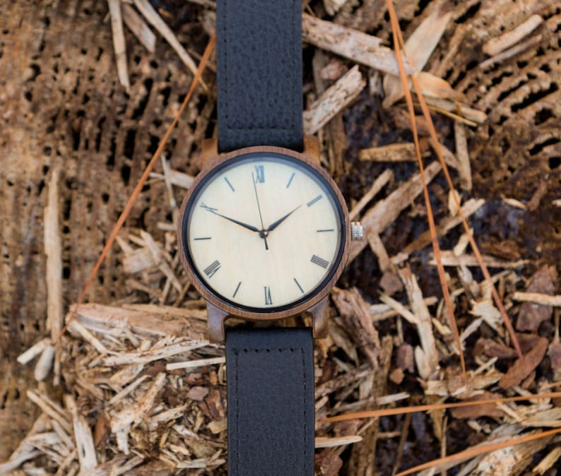 Pu&ograve; includere: Un orologio da polso in legno con un cinturino in pelle nera. Il quadrante dell'orologio &egrave; bianco con indici orari in numeri romani neri e lancette nere.