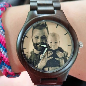 May include: A dark brown wooden wristwatch with a custom photo on the watch face. The watch face features a black and white image of a man holding a baby. The watch has a dark brown band and silver accents.