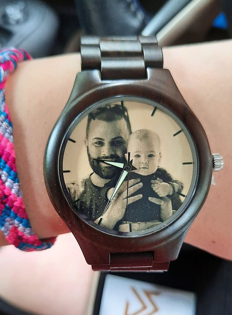 May include: A dark brown wooden wristwatch with a custom photo on the watch face. The watch face features a black and white image of a man holding a baby. The watch has a dark brown band and silver accents.