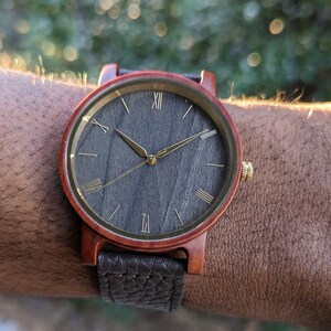 May include: A wooden wristwatch with a dark grey face and gold Roman numeral hour markers. The watch has a brown leather strap.