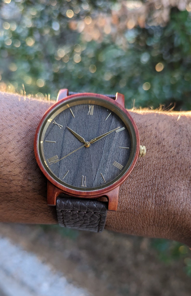 May include: A wooden wristwatch with a dark grey face and gold Roman numeral hour markers. The watch has a brown leather strap.