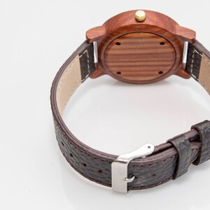 May include: A brown leather wristwatch with a wooden face and a gold-coloured knob. The watch has a brown leather strap with a silver buckle.