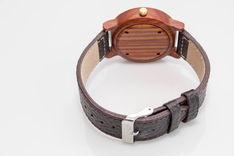 May include: A brown leather wristwatch with a wooden face and a gold-coloured knob. The watch has a brown leather strap with a silver buckle.
