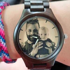 May include: A dark brown wooden wristwatch with a custom photo on the watch face. The watch face shows a black and white image of a man holding a baby. The watch has a dark brown strap and silver accents.