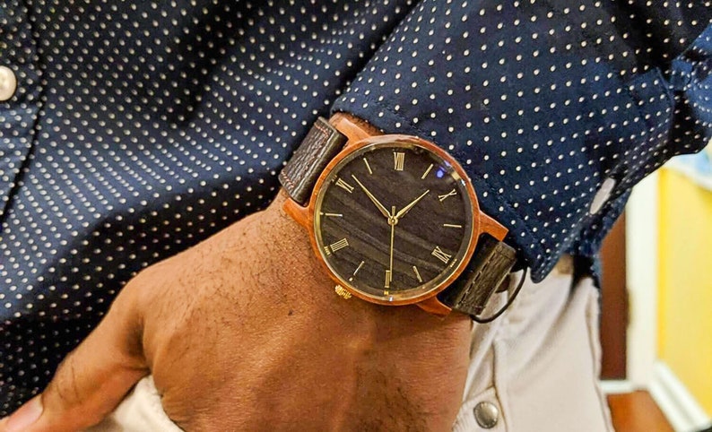 May include: A brown wooden wristwatch with a black face and gold Roman numeral hour markers. The watch has a brown leather strap.