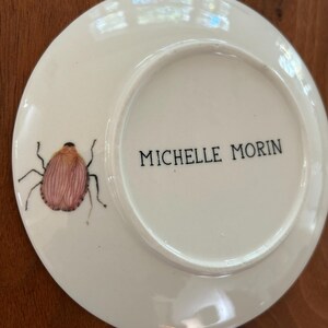 以下が含まれることがあります： ピンクと茶色の昆虫のイラストが描かれた白いセラミックプレート。プレートには黒で「MICHELLE MORIN」の文字が印刷されています。このプレートは、装飾用またはサービング用のアイテムである可能性があります。
