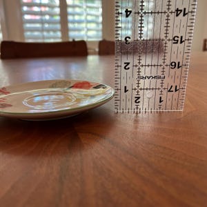 以下が含まれることがあります： 赤、黄、緑の花柄が施された、小さくて丸いベージュ色のセラミックソーサー。ソーサーは緑色の縁とくぼんだ中央部があります。ソーサーの隣には定規があり、約10cmの測定値を示しています。