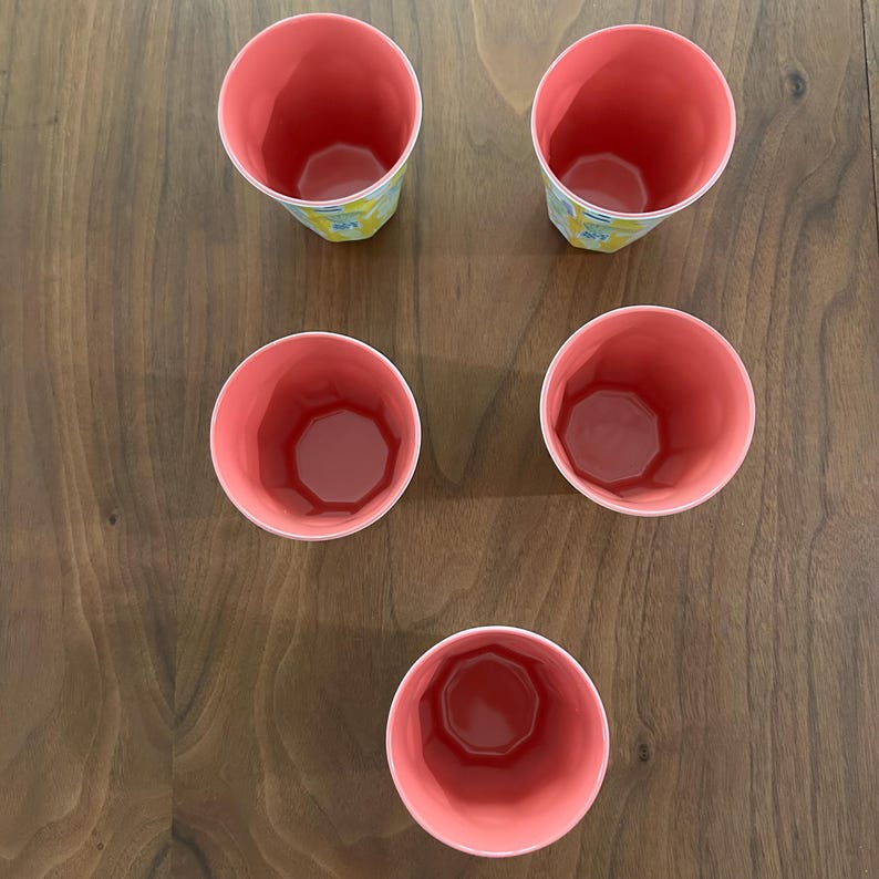 May include: Overhead view of five pink plastic cups. The cups have a geometric design on the outside and a solid pink interior. The cups are arranged on a wooden surface.