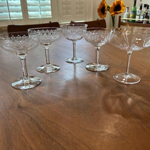 May include: Five clear, stemmed champagne coupe glasses with various etched designs are arranged on a dark wooden table. The glasses have round bases and delicate stems, showcasing intricate patterns and shapes.