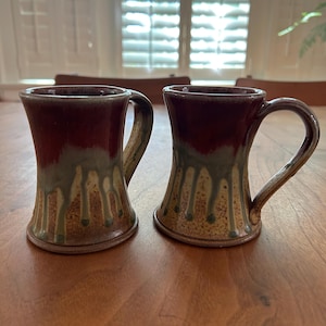 May include: Two handmade ceramic mugs with a unique design. The mugs feature a dark red top that fades into a green and tan base with a drip effect. Each mug has a curved handle and sits on a wooden table.