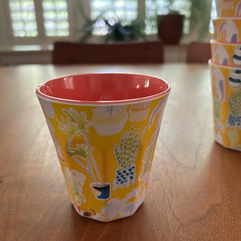 May include: A yellow cup with a coral interior and a botanical pattern of potted plants. The cup has a faceted design and is part of a set. The background is a wooden table with a blurred view of a window.